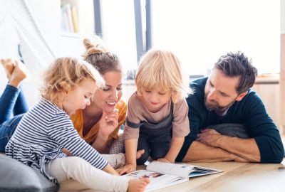 Front view of young family with two small children indoors in bedroom reading a book.